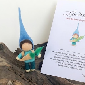 Pode incluir: Uma figura de gnomo de feltro azul, segurando uma cenoura e uma folha verde, ao lado de um cartão com texto em alemão e uma ilustração de gnomo.