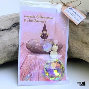 May include: A clear plastic-wrapped card featuring a gnome illustration and the text "magische Lichtmomente f&uuml;r dein Zuhause". A hanging ornament with a faceted crystal, a pale pink bead, and a wooden bead is attached. A tag reads "Echter Rosenquarz".