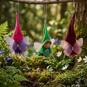 May include: Three whimsical fairy ornaments hanging from a branch. Each fairy has a pointed hat, butterfly wings, and a small wooden body. The fairies are in shades of red, green, and purple, with a forest background.