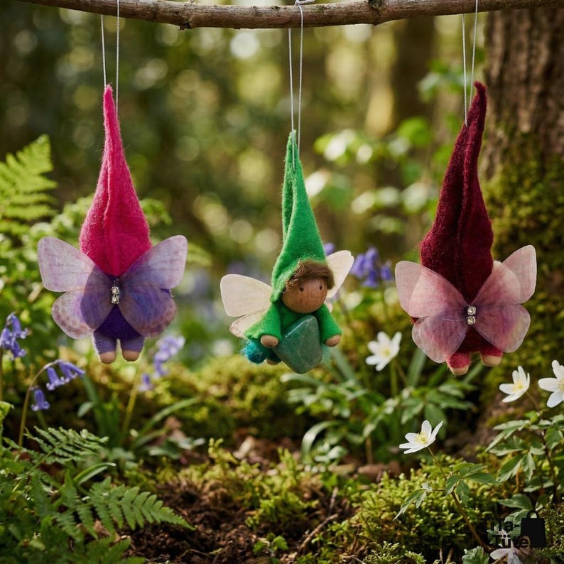 May include: Three whimsical fairy ornaments hanging from a branch. Each fairy has a pointed hat, butterfly wings, and a small wooden body. The fairies are in shades of red, green, and purple, with a forest background.