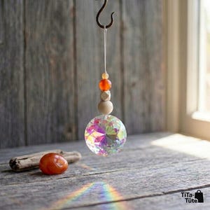 May include: A hanging suncatcher featuring a faceted clear crystal sphere that reflects iridescent colours. Above the sphere are a light wooden bead, a small square wooden bead, and a round orange stone bead, all suspended by a white string.