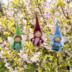 May include: Three handmade fairy ornaments in green, burgundy, and blue, each with a pointed hat, wooden face, and delicate wings. They are suspended from a tree branch with pink blossoms, creating a whimsical and charming scene.