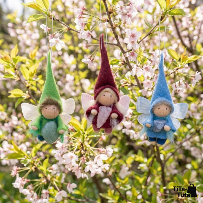 May include: Three handmade fairy ornaments in green, burgundy, and blue, each with a pointed hat, wooden face, and delicate wings. They are suspended from a tree branch with pink blossoms, creating a whimsical and charming scene.