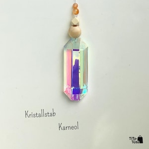 May include: A hanging ornament featuring a faceted, iridescent glass crystal prism suspended by a string with a round wooden bead, a cube-shaped wooden bead, and two small orange carnelian beads. The text Kristallstab Karneol and TiTa-T&uuml;te appear.