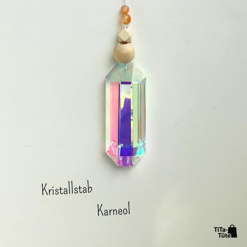 May include: A hanging ornament featuring a faceted, iridescent glass crystal prism suspended by a string with a round wooden bead, a cube-shaped wooden bead, and two small orange carnelian beads. The text Kristallstab Karneol and TiTa-T&uuml;te appear.