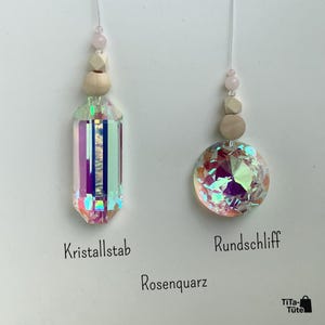 May include: Two hanging ornaments featuring iridescent, multi-faceted crystals suspended by strings. Each has a small pink bead and two light-coloured wooden beads above the crystal. Text labels read Kristallstab, Rundschliff, and Rosenquarz.