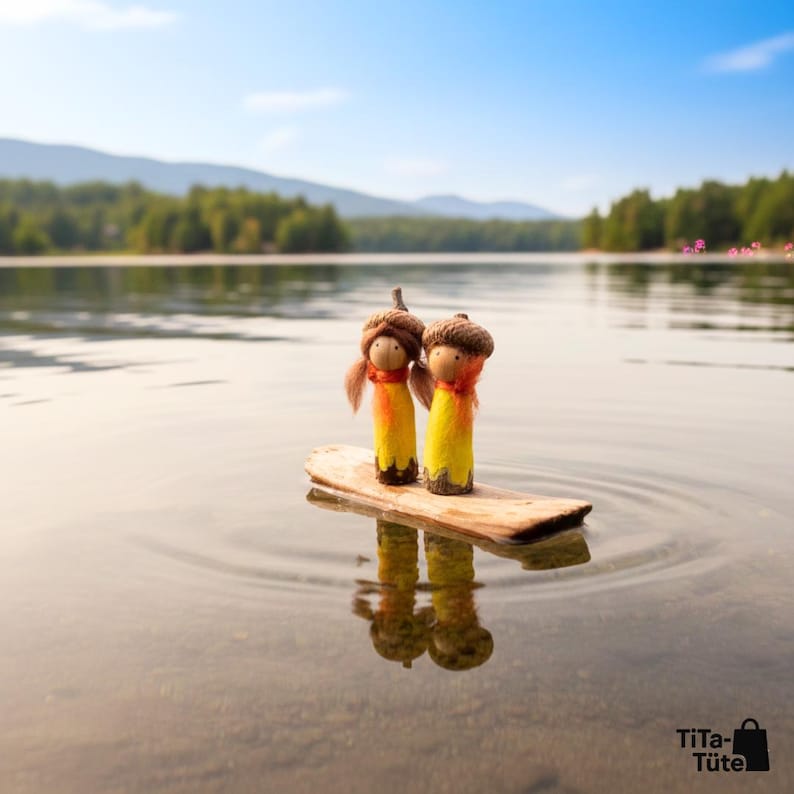 Idyllische Szene am See: Waldwichtel Pärchen 'Sonnenstrahl' treibt auf einem kleinen Holzfloß über das ruhige Wasser. Die gelben Figuren spiegeln sich sanft im See vor einer unscharfen Waldkulisse. Sommerliche Waldorf Deko und Symbol für die Reise des Lebens von TiTa-Tüte