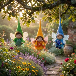 May include: Three handmade fairy dolls hanging from a tree branch. Each doll has a pointed hat and wings, with different colored outfits: green, yellow, and blue. The dolls are suspended in front of a garden with flowers and a stone path.