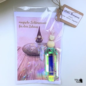May include: A clear plastic bag containing a rectangular card with a drawing of a gnome on a stone and the text "magische Lichtmomente f&uuml;r dein Zuhause". Attached is a hanging ornament with a large iridescent crystal, wooden beads, and a tag.