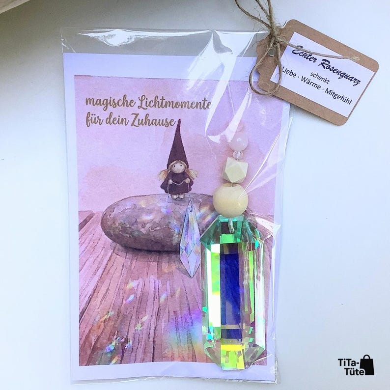 May include: A clear plastic bag containing a rectangular card with a drawing of a gnome on a stone and the text "magische Lichtmomente f&uuml;r dein Zuhause". Attached is a hanging ornament with a large iridescent crystal, wooden beads, and a tag.