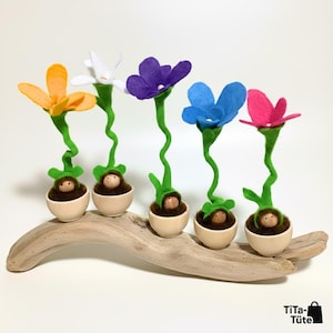 Könnte beinhalten: Fünf Filzblumenskulpturen in Orange, Weiß, Lila, Blau und Rosa, jede mit einem grünen Stiel und einer kleinen Figur in einem Holztopf. Die Blumen sind auf einem Stück Treibholz angeordnet.