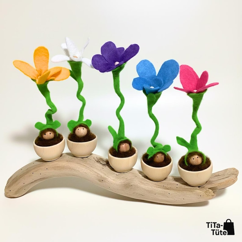 Fünf bunte Filz-Blumenfiguren von TiTa-Tüte balancieren auf einem Stück Treibholz. Minimalistische Präsentation der Wurzel-Wichtel vor weißem Hintergrund, ideal um die geschwungenen grünen Stiele zu zeigen