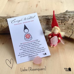 Op de afbeelding: Een handgemaakte pop met een rode hoed en jurk, die een roze rozenkwartskristal vasthoudt, naast een kaart met Duitse tekst. De kaart toont een tekening van een figuur en de woorden "Energie Wichtel" en "Liebe (Rosenquarz)".