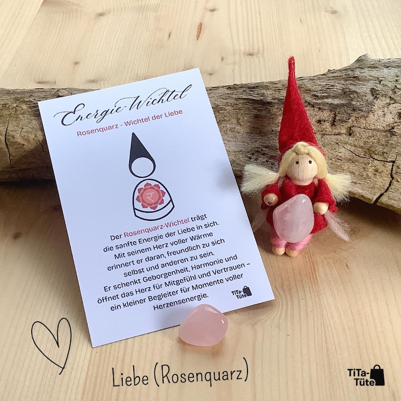 Peut inclure: Une poup&eacute;e artisanale avec un chapeau et une robe rouges, tenant un cristal de quartz rose, &agrave; c&ocirc;t&eacute; d'une carte avec du texte allemand. La carte pr&eacute;sente un dessin d'une figurine et les mots "Energie Wichtel" et "Liebe (Rosenquarz)".