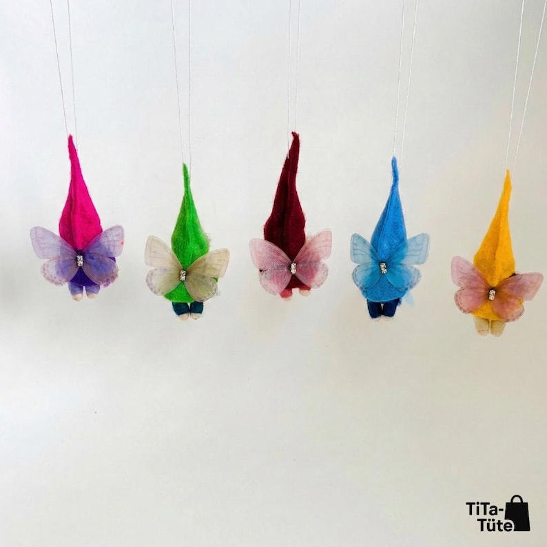 May include: Five whimsical gnome ornaments with colorful pointed hats and delicate butterfly wings. Each gnome features a different hat color: pink, green, burgundy, blue, and yellow. The wings are adorned with a small jewel. The ornaments are suspended against a white background.