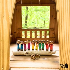 Die bunte Waldwichtel-Familie im hölzernen Spielhaus: Alle Figuren der Kollektion stehen aufgereiht auf einem Treibholz-Ast vor dem Fenster mit Blick in den grünen Wald. Handgeschnitztes Waldorf Spielzeug in Regenbogenfarben von TiTa-Tüte.