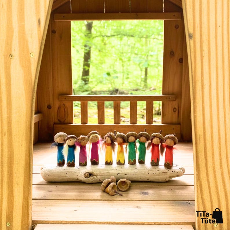 Die bunte Waldwichtel-Familie im hölzernen Spielhaus: Alle Figuren der Kollektion stehen aufgereiht auf einem Treibholz-Ast vor dem Fenster mit Blick in den grünen Wald. Handgeschnitztes Waldorf Spielzeug in Regenbogenfarben von TiTa-Tüte.