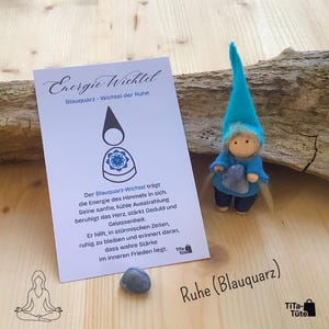 Könnte beinhalten: Eine handgefertigte Zwergenpuppe aus Filz mit blauem Hut und Outfit, die einen blauen Kristall hält. Eine Karte mit deutschem Text, "Energie-Wichtel" und "Ruhe (Blauquarz)", liegt neben der Puppe. Auf Holzuntergrund.