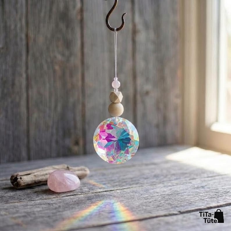 May include: A hanging suncatcher featuring a large, multifaceted iridescent glass sphere suspended by a thin string. Above the sphere are a small light pink bead and a larger, smooth, light-coloured wooden bead. A polished rose quartz stone sits nearby.