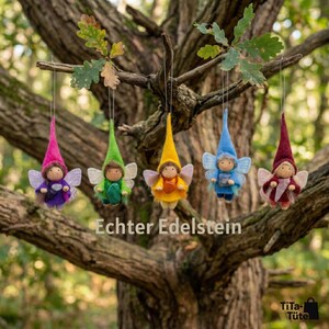 May include: Five colorful felt fairy ornaments hanging from a tree branch. Each fairy has a pointed hat, wings, and a small body. The fairies are in shades of pink, green, yellow, blue, and burgundy. The text "Echter Edelstein" is visible.