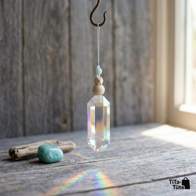 May include: A hanging suncatcher ornament featuring a clear, iridescent, multi-faceted crystal prism suspended by a white string. Above the prism are two round wooden beads and one small light green bead. A smooth, rounded green stone sits below.