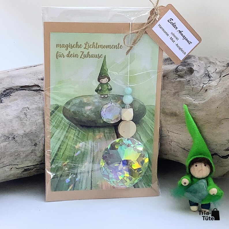 May include: A suncatcher ornament featuring a large faceted crystal, light blue beads, and wooden spheres, packaged with a card displaying a gnome illustration and the text "magische Lichtmomente f&uuml;r dein Zuhause". Includes a tag labelled "Echter Amazonit".