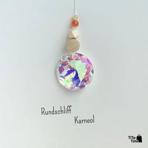 May include: A hanging ornament featuring a large, circular, iridescent crystal prism suspended below a light-coloured wooden bead, a faceted wooden bead, and two small orange beads. The text "Rundschliff Karneol" is printed below the ornament.