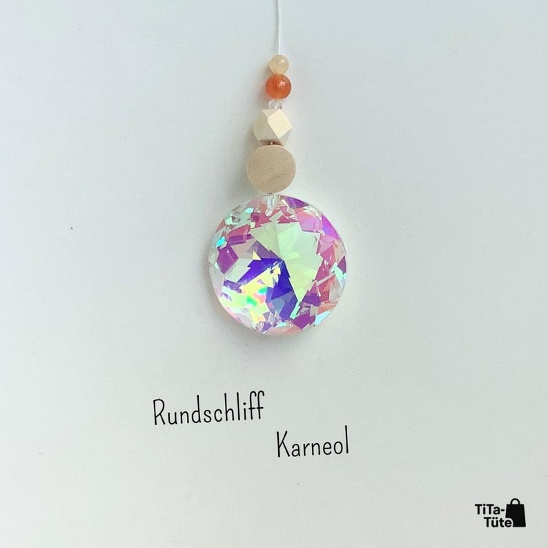 May include: A hanging ornament featuring a large, circular, iridescent crystal prism suspended below a light-coloured wooden bead, a faceted wooden bead, and two small orange beads. The text "Rundschliff Karneol" is printed below the ornament.