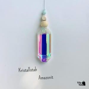 May include: A hanging ornament featuring a faceted, iridescent crystal prism with a rainbow sheen. Above the crystal are two light-coloured wooden beads and a small, pale blue stone bead. The text Kristallstab Amazonit is visible below.
