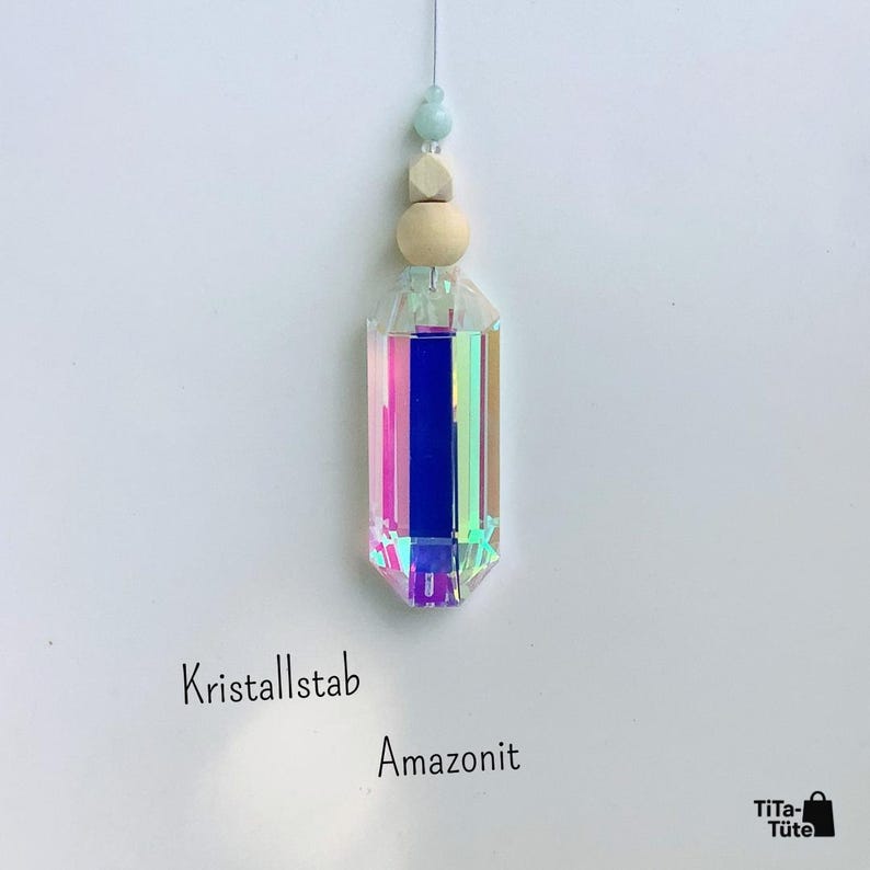 May include: A hanging ornament featuring a faceted, iridescent crystal prism with a rainbow sheen. Above the crystal are two light-coloured wooden beads and a small, pale blue stone bead. The text Kristallstab Amazonit is visible below.