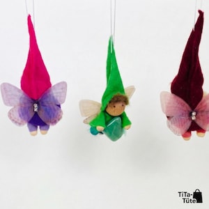 May include: Three whimsical fairy ornaments hanging against a white background. Each fairy has a pointed hat, butterfly wings, and a small wooden body. The hats are in shades of pink, green, and burgundy. The wings are translucent with delicate patterns.