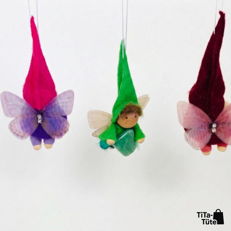 May include: Three whimsical fairy ornaments hanging against a white background. Each fairy has a pointed hat, butterfly wings, and a small wooden body. The hats are in shades of pink, green, and burgundy. The wings are translucent with delicate patterns.