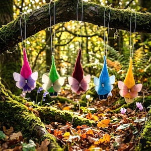 May include: Five whimsical felt gnome ornaments with butterfly wings hang from a tree branch. Each gnome has a different colored pointed hat: pink, green, burgundy, blue, and yellow. The wings are sheer and adorned with a small gem.