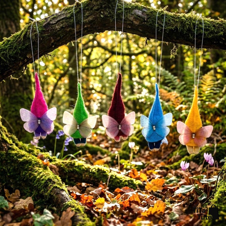 May include: Five whimsical felt gnome ornaments with butterfly wings hang from a tree branch. Each gnome has a different colored pointed hat: pink, green, burgundy, blue, and yellow. The wings are sheer and adorned with a small gem.