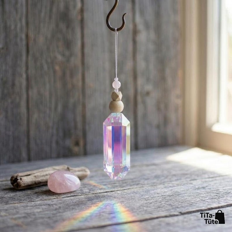 May include: A hanging iridescent crystal prism ornament suspended by a white string from a metal hook. The prism features a faceted hexagonal shape with rainbow light reflections, topped with a small pink bead and a round wooden bead.