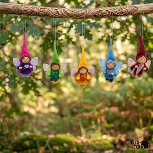May include: Five handmade fairy ornaments with pointed hats and wings hang from a carved wooden branch. Each fairy is dressed in a different color: pink, green, yellow, blue, and burgundy. The background is a blurred forest scene.