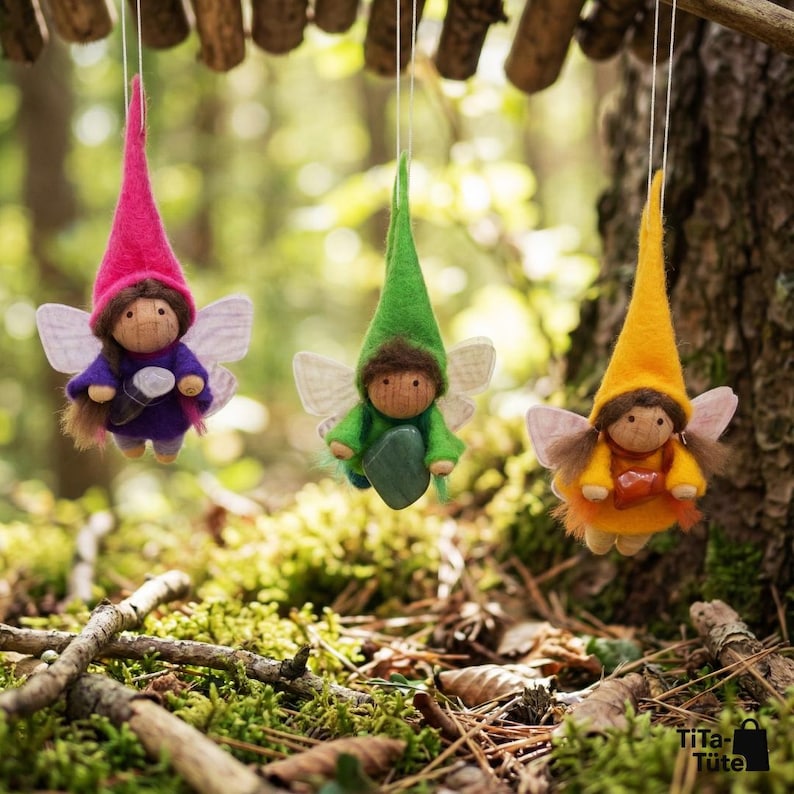 May include: Three handmade fairy dolls with felt hats and wings hang from a wooden branch. The dolls have wooden faces and are dressed in purple, green, and yellow outfits. The background is a blurred forest scene with green moss and tree trunks.