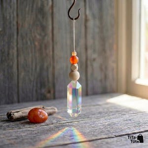 May include: A hanging ornament featuring a clear, iridescent, hexagonal crystal prism suspended by a white string. Above the prism are a round wooden bead, a faceted wooden bead, and a small orange bead. A smooth orange stone sits nearby.