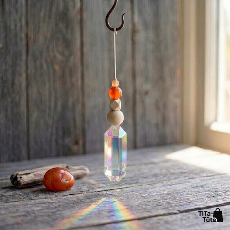 May include: A hanging ornament featuring a clear, iridescent, hexagonal crystal prism suspended by a white string. Above the prism are a round wooden bead, a faceted wooden bead, and a small orange bead. A smooth orange stone sits nearby.