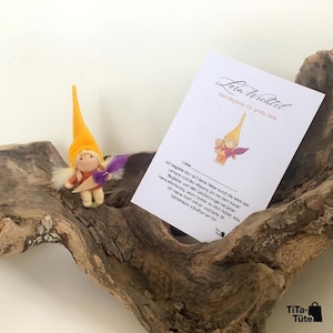 May include: A handmade felt gnome figurine with a yellow hat, purple wings, and a small card. The card has text in German and an illustration of a gnome. The gnome is on a piece of driftwood.