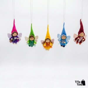 May include: Five hanging fairy ornaments with colorful pointed hats and wings. Each fairy has a different outfit: purple, green, yellow, blue, and burgundy. The fairies are suspended by thin strings against a white background.