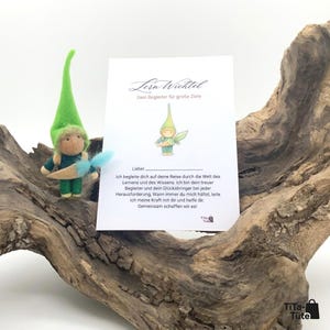 Könnte beinhalten: Eine handgefertigte Filzpuppe mit grünem Hut und blauen Akzenten, neben einer Karte mit deutschem Text. Die Puppe befindet sich auf einem Stück Treibholz. Auf der Karte steht "Lern-Wichtel Dein Begleiter für große Ziele".