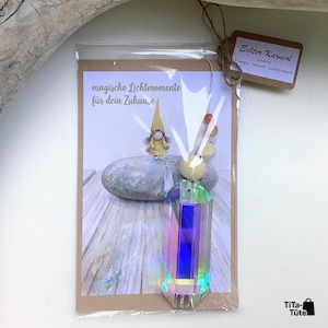 May include: A clear, iridescent, rectangular glass prism pendant attached to a card in a plastic sleeve. The card features a drawing of a gnome on a stone with the text 'magische Lichtmomente f&uuml;r dein Zuhause'. A brown tag reads 'Echter Karneol'.