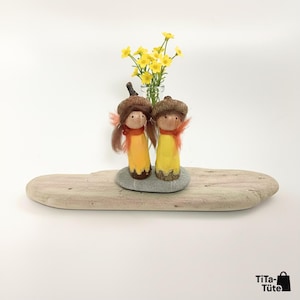 Handgeschnitztes Waldwichtel Pärchen 'Sonnenstrahl' in Gelb, arrangiert auf einem Stück Treibholz und einem Kieselstein. Im Hintergrund steht eine kleine Vase mit gelben Wiesenblumen. Fröhliche Waldorf Frühlings- und Sommerdeko von TiTa-Tüte.
