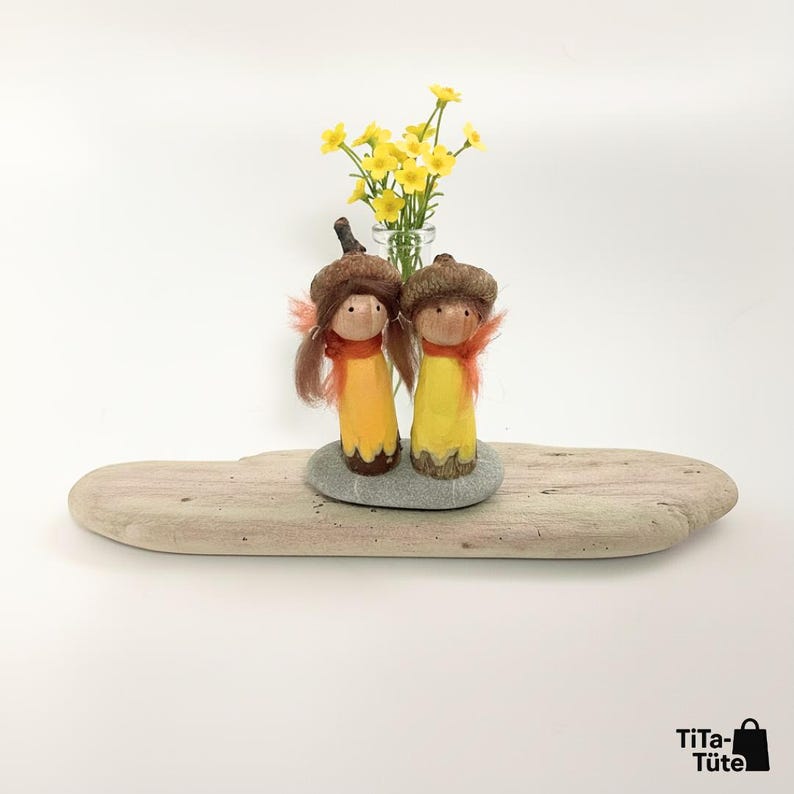 Handgeschnitztes Waldwichtel Pärchen 'Sonnenstrahl' in Gelb, arrangiert auf einem Stück Treibholz und einem Kieselstein. Im Hintergrund steht eine kleine Vase mit gelben Wiesenblumen. Fröhliche Waldorf Frühlings- und Sommerdeko von TiTa-Tüte.