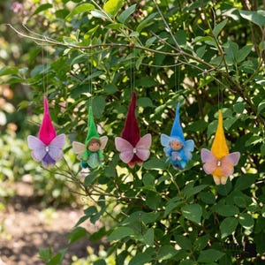 May include: Five whimsical fairy ornaments with colorful pointed hats and butterfly wings hang from a tree branch. Each fairy has a different colored hat: pink, green, burgundy, blue, and yellow. The wings are sheer and adorned with a small jewel.
