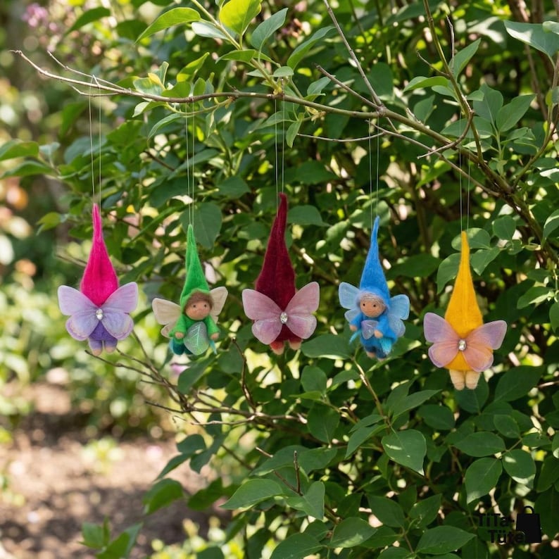 May include: Five whimsical fairy ornaments with colorful pointed hats and butterfly wings hang from a tree branch. Each fairy has a different colored hat: pink, green, burgundy, blue, and yellow. The wings are sheer and adorned with a small jewel.