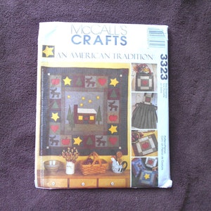 Könnte beinhalten: McCall's Crafts Nähmuster-Umschlag mit einem rot-weiß-blauen Farbschema. Das Muster ist für eine Steppdecke mit einer Hütte, Bäumen, Sternen und Herzen. Die Musternummer ist 3323. Der Text "An American Tradition" ist auf dem Umschlag gedruckt.