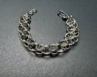 May include: A silver-colored chainmail bracelet with a hook clasp. The bracelet is made of small, interlocking metal rings, creating a textured, woven appearance. The jewelry is displayed on a dark gray surface.