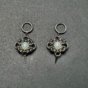 Puede incluir: Un par de pendientes plateados. Cada pendiente presenta una parte superior circular, un diseño decorativo tejido y una piedra central ovalada e iridiscente. Una pequeña punta puntiaguda cuelga de la parte inferior de cada pendiente.